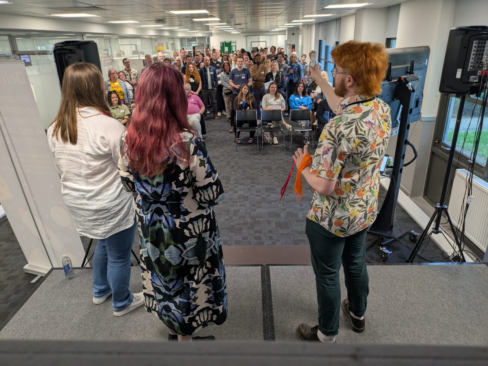 Three people on stage speaking speaking to a crown of 100 people within a meeting room