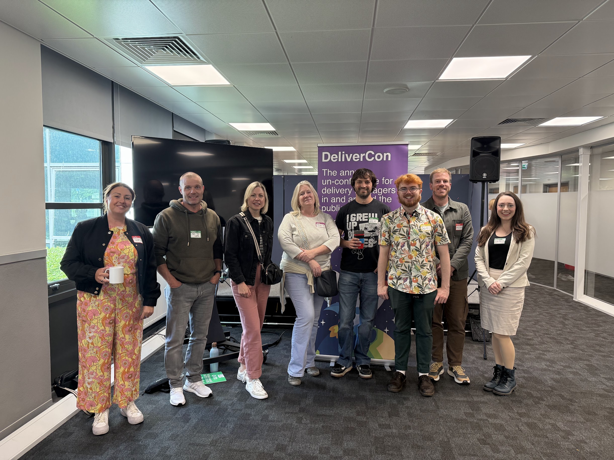 Eight smiling people standing in frount of a DeliverCon Sign
