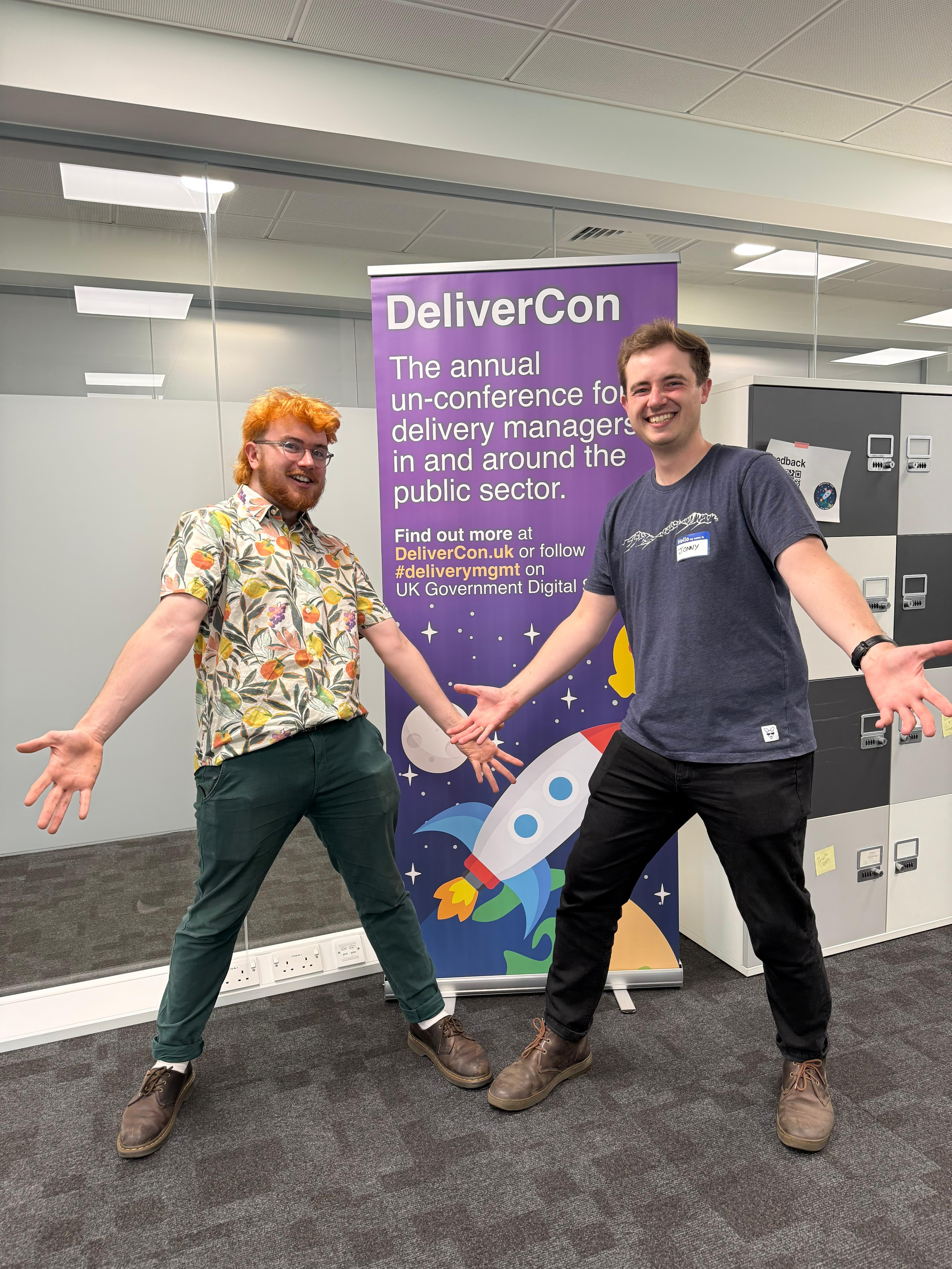 Two men standing in fornt of the DeliverCon Banner with hands out in an exaggerated pose.